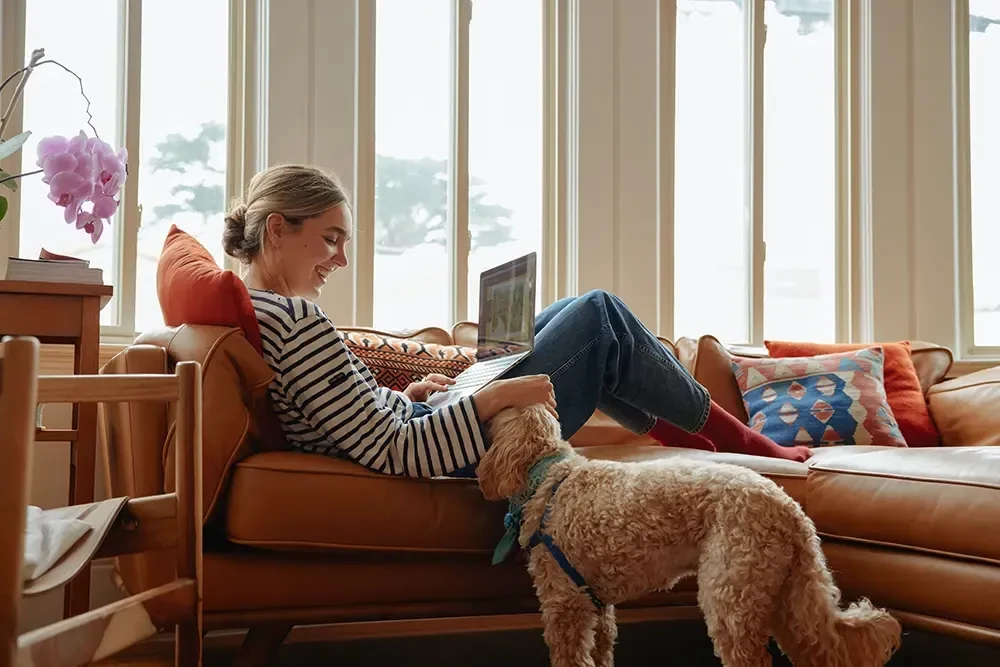 Femme assise sur un canapé avec un chien à ses pieds, travaillant sur un ordinateur portable près d'une fenêtre lumineuse.